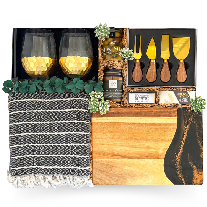 Luxury entertaining gift basket in a large high-gloss black magnetic keepsake gift box featuring premium gold stemless wine glasses, artisan serving board, cheese knife set, gourmet foods, cozy throw, candle, and elegant home entertaining essentials.

