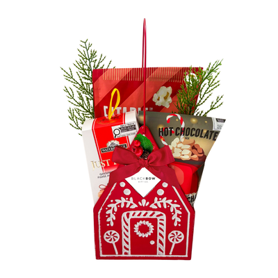 A Cozy Christmas holiday gift featuring a red gingerbread house keepsake filled with festive popcorn, hot chocolate mix, gingerbread fudge and seasonal greenery.
