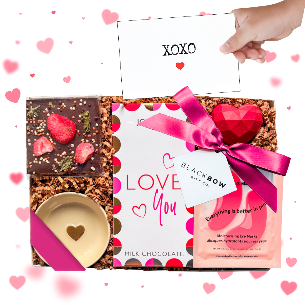 A clear-lid gift box tied with a bright pink satin ribbon and accented with delicate details, displaying a strawberry mint chocolate bar, red heart-shaped lip balm, small gold heart ceramic dish, pink moisturizing under-eye mask packet, and a heart pick accent—styled in a romantic pink and gold palette for a modern, thoughtful gifting presentation.