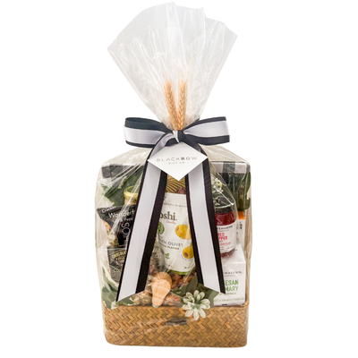 Woven picnic gift basket featuring a bamboo serving board, tea towels, gourmet snacks, olives, pistachios, popcorn, chocolates, and entertaining essentials by Black Bow Gift Co.
