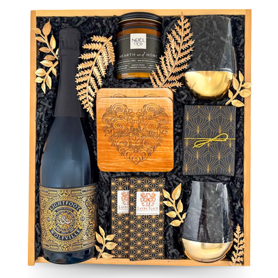 Luxury wooden keepsake gift box featuring premium sparkling wine, two gold-accented stemless wine glasses, artisan chocolate, scented candle, engraved wooden coaster, gourmet treats, and elegant celebration-inspired gifts.