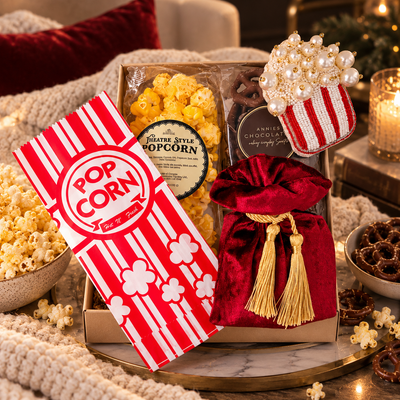 Luxury movie night gift set featuring gourmet popcorn, chocolate-covered pretzels, a red velvet pouch with gold tassels, and a beaded popcorn ornament in an elegant, cozy living room setting.