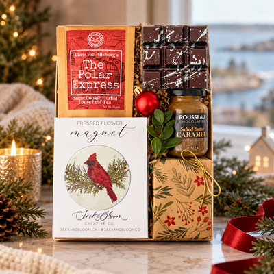 Luxury Nova Scotia-inspired holiday gift box featuring Polar Express tea, artisan chocolate, salted butter caramel, and a cardinal keepsake, styled with evergreens, candlelight, and an elegant coastal Christmas backdrop.