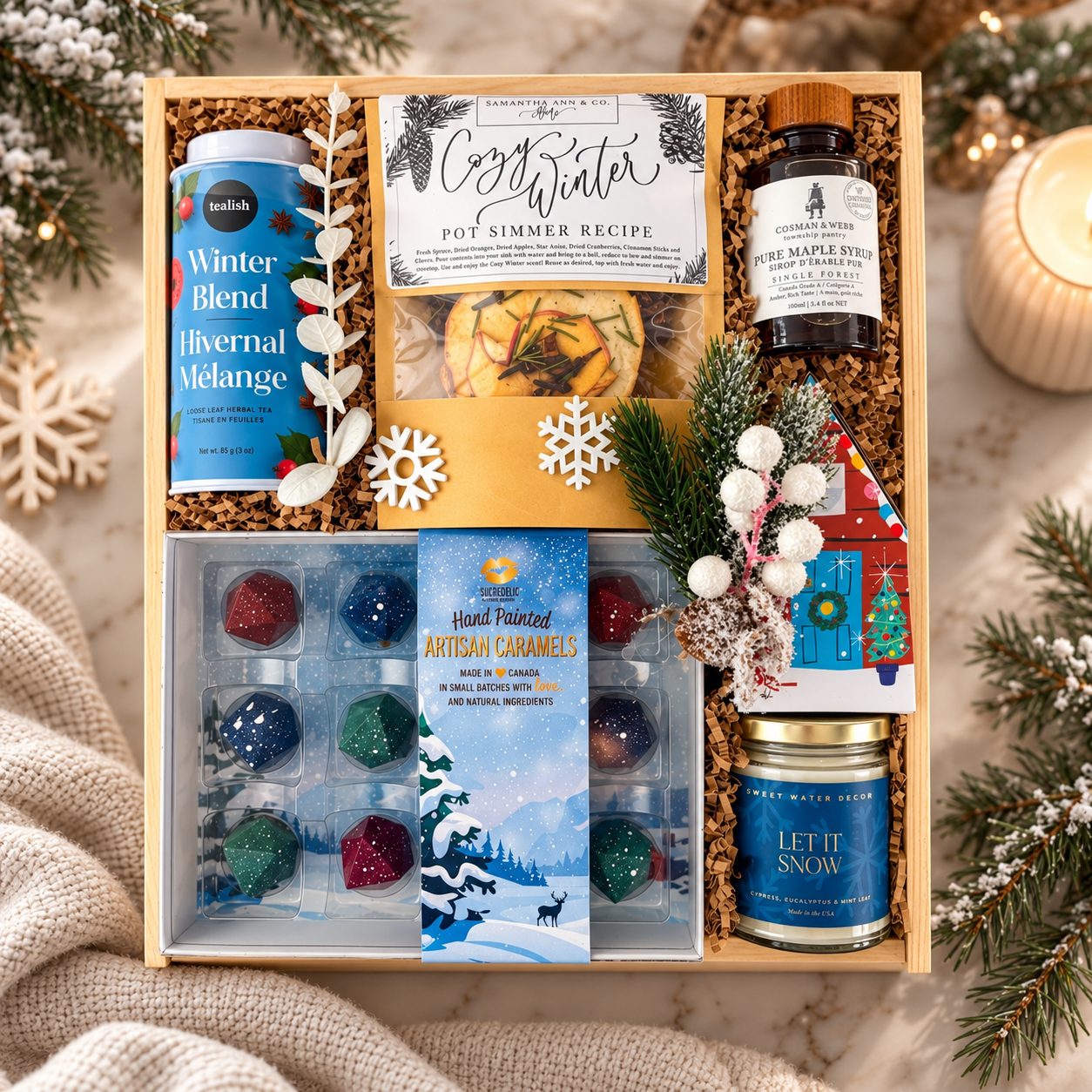 Luxury winter gift box featuring Winter Blend tea, maple syrup, artisan caramels, cozy simmer pot kit and Let It Snow candle, elegantly styled with snowy greenery, soft knit textures and warm winter accents.