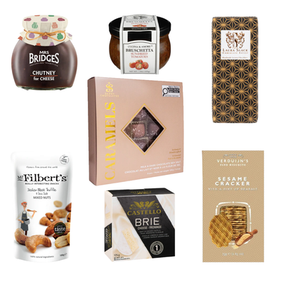 A flat-lay arrangement of gourmet food products against a white background. The items include Mrs. Bridges Chutney for Cheese, Cucina & Amore Sun-Dried Tomato Bruschetta, Laura Slack artisan chocolate, a box of Sea Salt Chocolate covered Caramels, Mr. Filbert's Italian Black Truffle & Sea Salt Mixed Nuts, a box of Castello Brie Cheese, and Verduijn's Sesame Crackers.