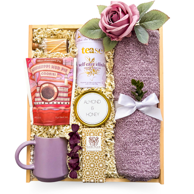 Elegant self-care gift featuring tea, chocolate, biscuits, candle, matches, mug, and a soft lilac scarf styled in a refined, calming palette.