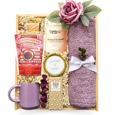 Elegant self-care gift featuring tea, chocolate, biscuits, candle, matches, mug, and a soft lilac scarf styled in a refined, calming palette.