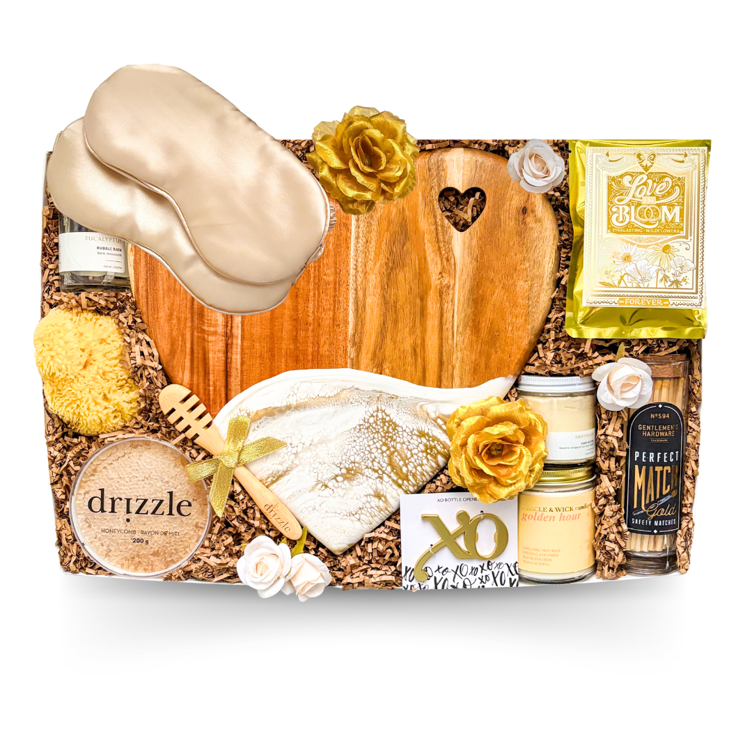 XL luxury keepsake gift box featuring a heart-shaped serving board, resin heart dish, silk sleep masks, bubble bath, candle, honey, sponge, bottle opener, safety matches, gourmet treats, olives, crackers, and charcuterie favourites.

