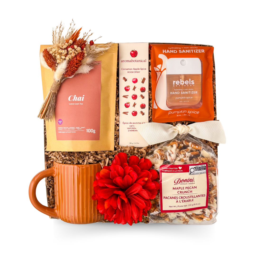 A curated gift basket set on a white background, featuring an orange ribbed ceramic mug, a bag of Chai tea, a box of aromabotanical cinnamon bark tea, a bag of Rebels Hand Sanitizer, a bag of Danili maple crunch snack mix, shredded paper filler, a large red flower, and a dried floral sprig tied with a cream ribbon.