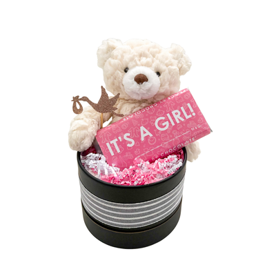 Keepsake baby girl gift with fluffy teddy bear and “It’s a Girl” chocolate, packaged in a round basket box.