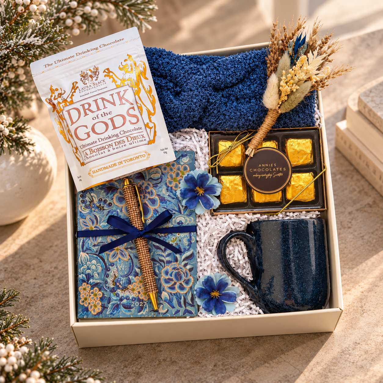Luxury winter gift box featuring drinking chocolate, gold-wrapped chocolates, a blue floral 2027 journal with pen, navy mug, cozy blue textile and dried flowers, elegantly styled in a warm, minimalist setting.