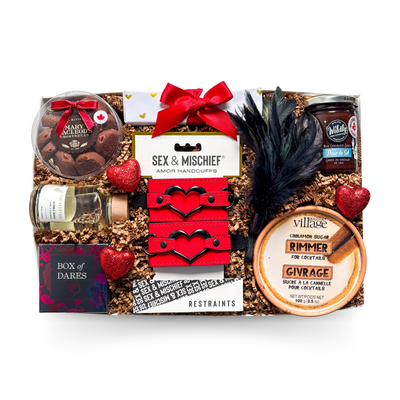 A black gift box tied with a satin ribbon and finished with heart accents, showcasing a playful date-night theme with chocolate treats, cinnamon-scented items, a eucalyptus bubble bath, a couples dare game, red plush handcuffs, a black feather, and a few cheeky extras for a bold and elevated gifting experience.