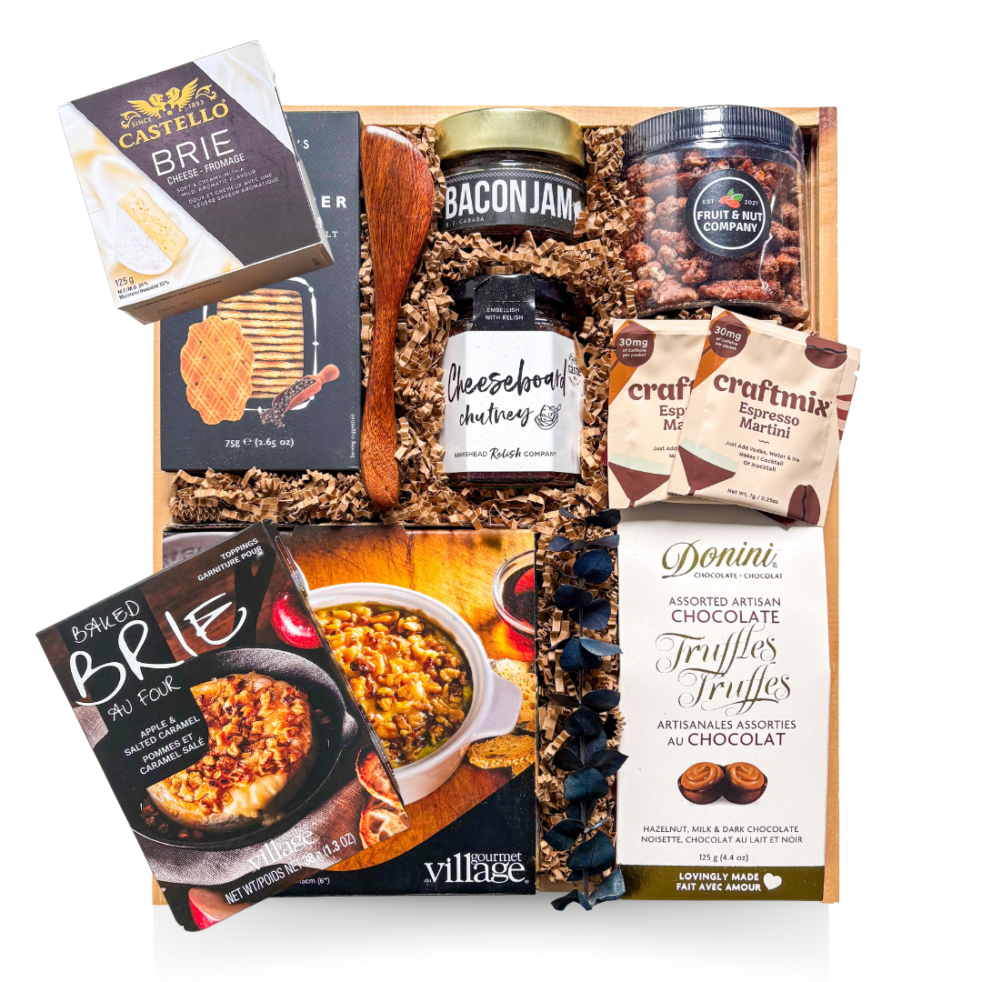 A premium wooden gift box tied with a grosgrain ribbon and accented with dried florals, filled with gourmet entertaining items including brie-themed accompaniments, crackers, spreads, nuts, cocktail mixers, chocolate truffles, and a white ceramic brie baker—styled for an elevated, shareable entertaining experience.