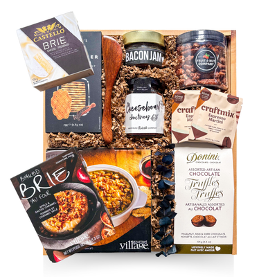 A premium wooden gift box tied with a grosgrain ribbon and accented with dried florals, filled with gourmet entertaining items including brie-themed accompaniments, crackers, spreads, nuts, cocktail mixers, chocolate truffles, and a white ceramic brie baker—styled for an elevated, shareable entertaining experience.