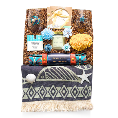An XL luxury gift box tied with a satin ribbon and accented with dried coastal florals, showcasing a seaside-themed assortment including stemless beach glassware, coastal coasters, a candle, natural sea sponge, gourmet shortbread, and a travel backgammon set—styled in soft, beachy tones for an elevated coastal gifting experience.