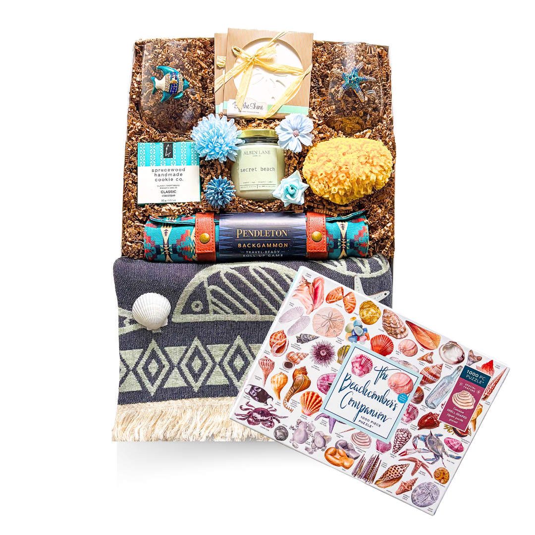 An XL luxury gift box tied with a satin ribbon and accented with dried coastal florals, showcasing a seaside-themed assortment including stemless beach glassware, coastal coasters, a candle, natural sea sponge, gourmet shortbread, and a travel backgammon set—styled in soft, beachy tones for an elevated coastal gifting experience.