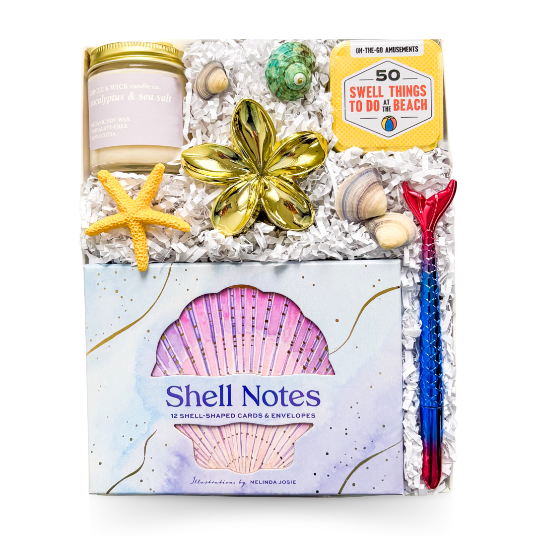 Luxury coastal gift box featuring shell stationery, coastal soy candle, beach activity cards, decorative seashells, floral trinket dish, mermaid pen, and seaside-inspired keepsakes.

