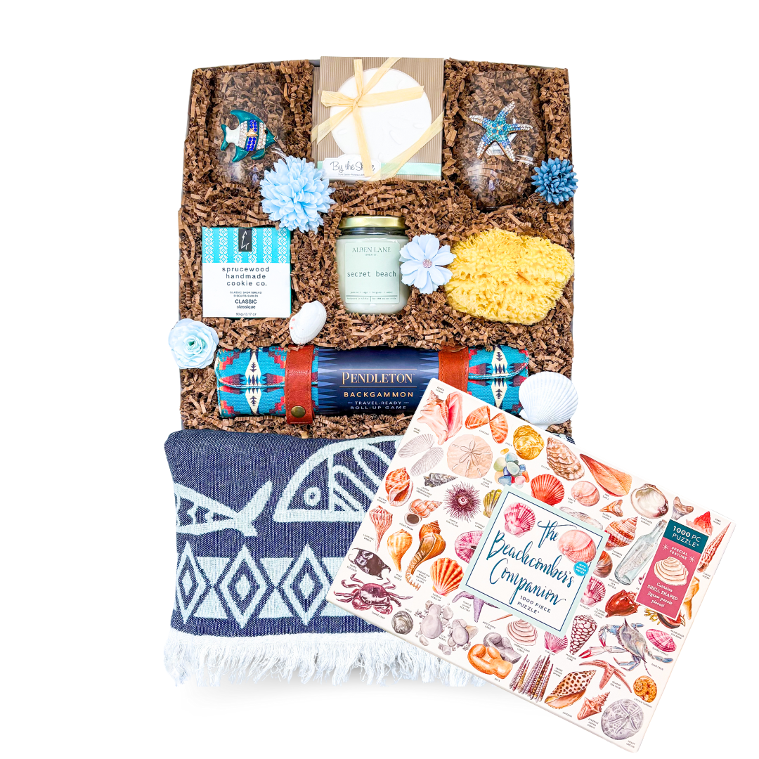 An XL luxury gift box tied with a satin ribbon and accented with dried coastal florals, showcasing a seaside-themed assortment including stemless beach glassware, coastal coasters, a candle, natural sea sponge, gourmet shortbread, and a travel backgammon set—styled in soft, beachy tones for an elevated coastal gifting experience.