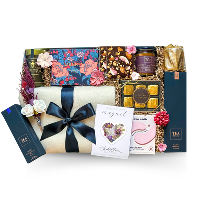The Art of Leisure luxury gift box featuring a soft cream throw blanket, floral playing cards, gourmet tea, chocolates, candle, coasters, pressed flower keepsake and self-care treats.