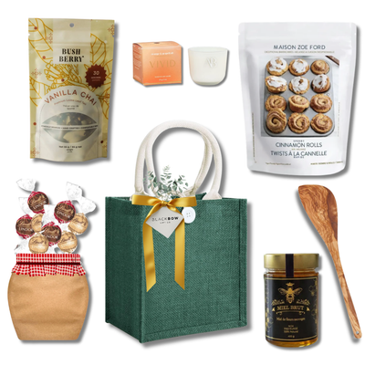 The Cozy Kitchen gift basket with cinnamon roll baking mix, vanilla chai, honey, chocolates, candle and wooden serving spoon presented in a reusable green tote by Black Bow Gift Co.