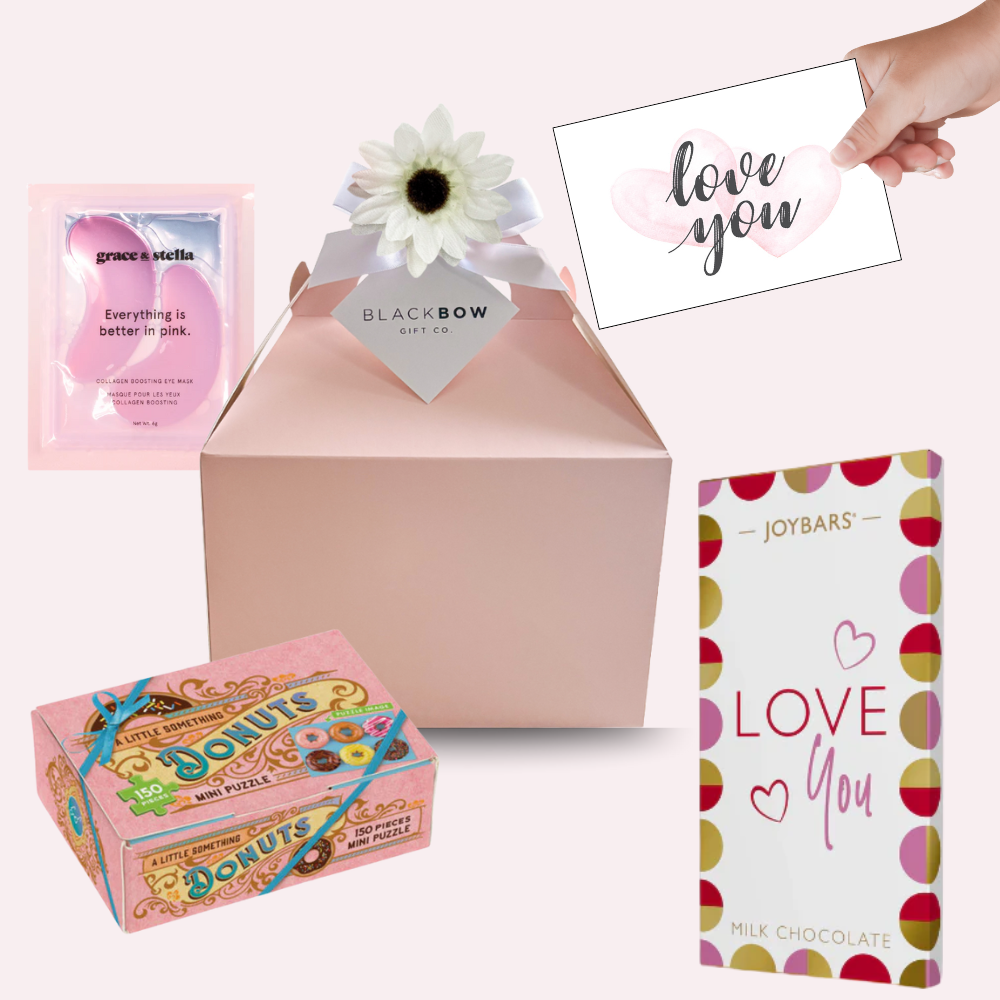 A blush pink gable gift box with a white ribbon and daisy flower topper, surrounded by a pink “Love You” milk chocolate bar, a pink collagen eye mask packet, and a small pastel donut-themed mini puzzle box—creating a sweet, playful, and self-care themed gift set.