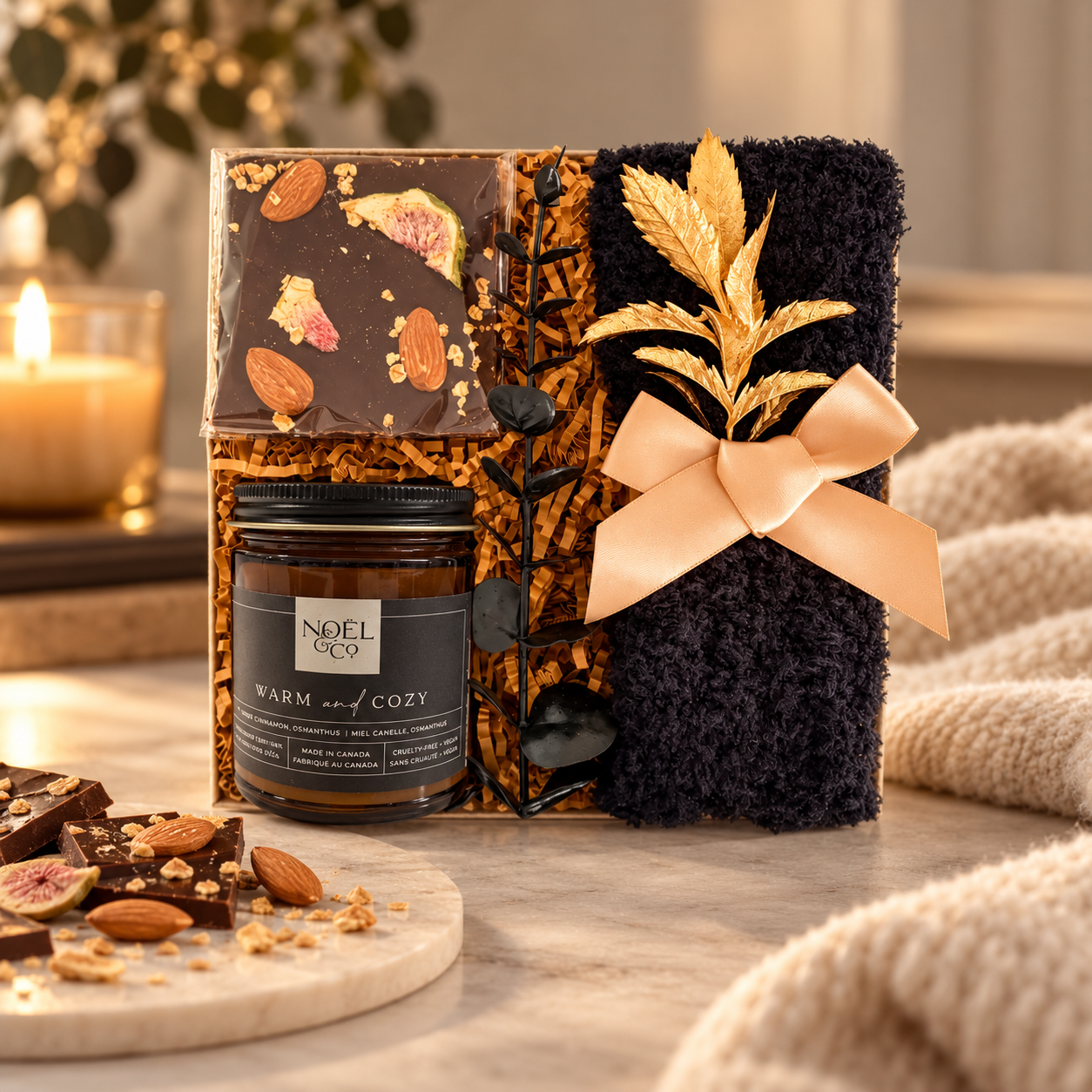 Luxury cozy gift box featuring artisan chocolate with almonds and figs, a Noël & Co. Warm and Cozy candle, and a plush black blanket with gold accents, styled in an elegant, warmly lit setting.