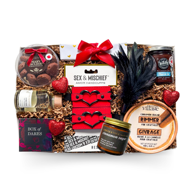 A black gift box tied with a satin ribbon and finished with heart accents, showcasing a playful date-night theme with chocolate treats, cinnamon-scented items, a eucalyptus bubble bath, a couples dare game, red plush handcuffs, a black feather, and a few cheeky extras for a bold and elevated gifting experience.