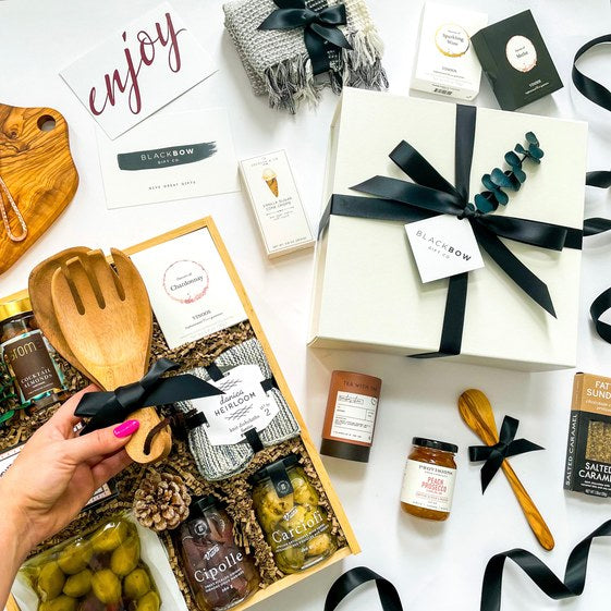 A personalised gift being assembled with a wooden board, kitchen utensils, gourmet treats and an enjoy card.