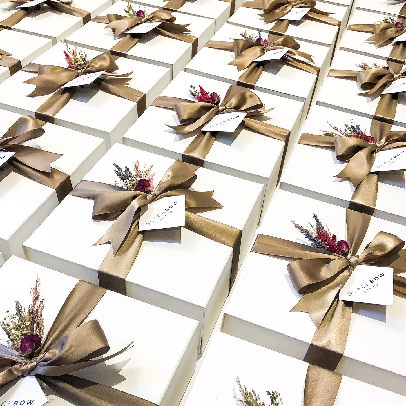 Rows of finished white gift boxes tied with taupe ribbon, a tag and dried flowers.